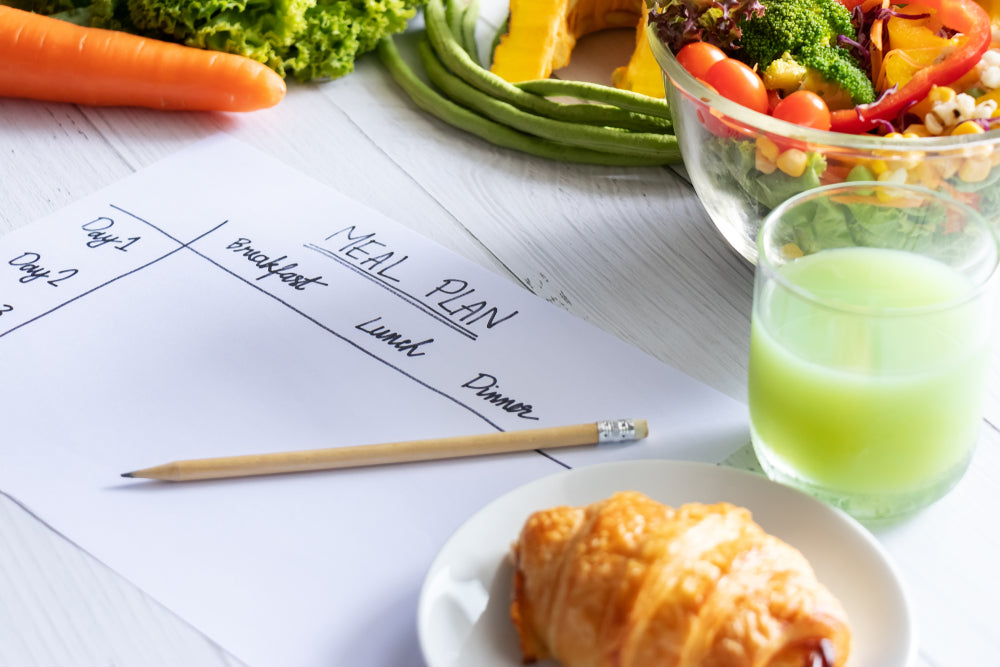 A meal planning sheet with sections for breakfast, lunch, and dinner sits on a table alongside fresh vegetables, a bowl of salad, a croissant, and a glass of green drink.