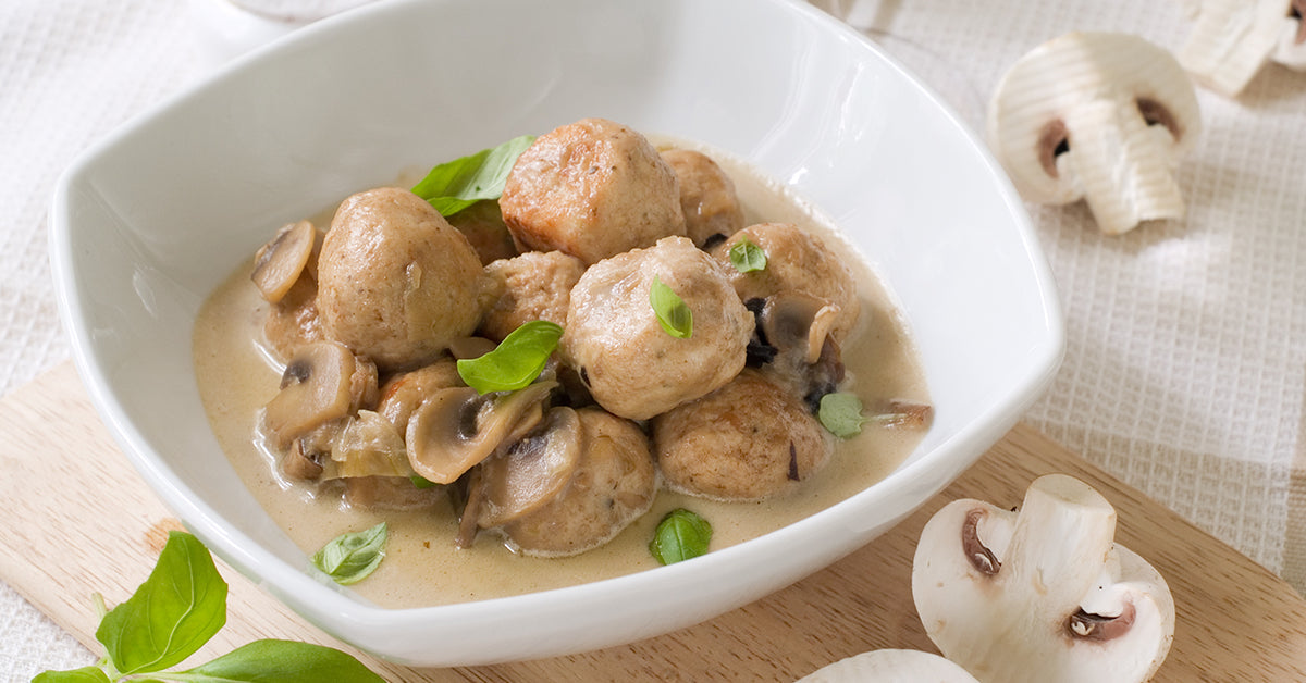 Bowl of meatballs in creamy mushroom sauce garnished with basil, with fresh mushrooms placed nearby.