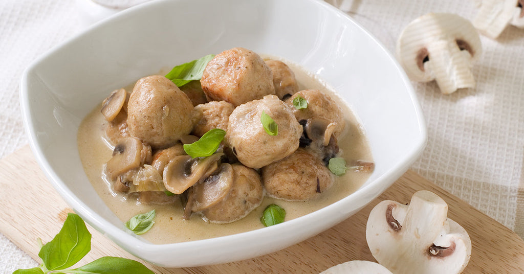 Bowl of meatballs in creamy mushroom sauce garnished with basil, with fresh mushrooms placed nearby.