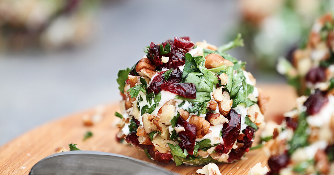 Cheese ball coated in chopped nuts, herbs, and dried cranberries on a wooden board.