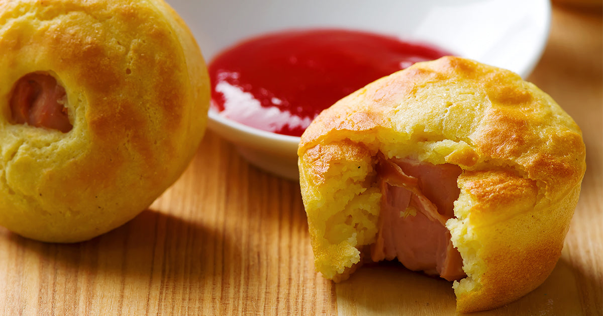 Two golden cornbread muffins, one partially bitten revealing a piece of hot dog inside, sit on a wooden surface next to a small bowl of ketchup.