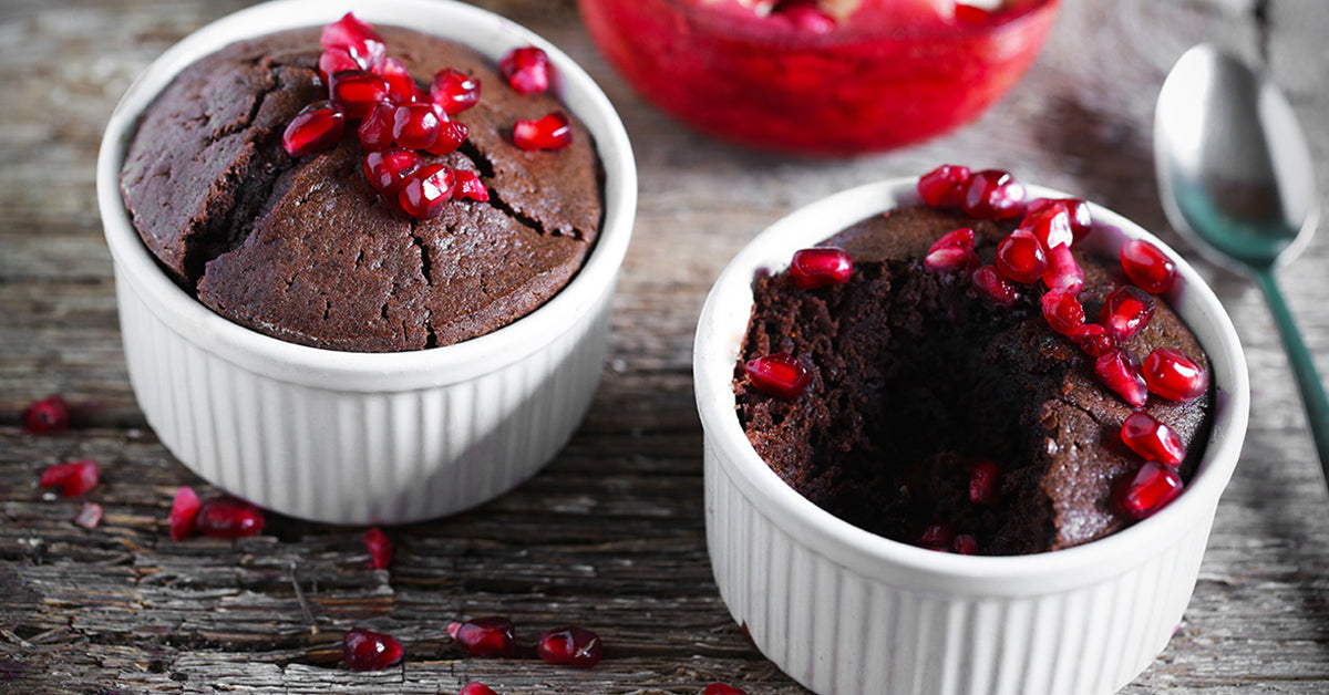 Chocolate lava cakes in white ramekins topped with pomegranate seeds on a rustic wooden surface.