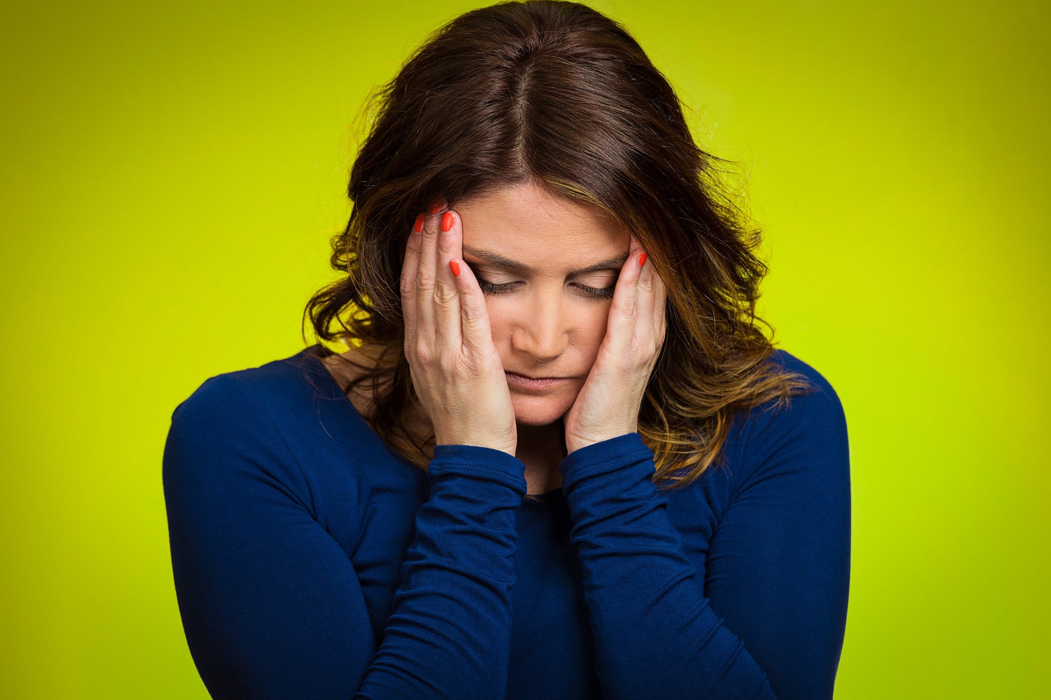 Woman in a blue shirt holding her face with both hands, looking down against a bright yellow background