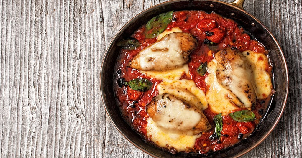 A skillet filled with three pieces of grilled chicken topped with melted cheese, resting in a rich tomato sauce with fresh basil leaves, set on a rustic wooden surface.