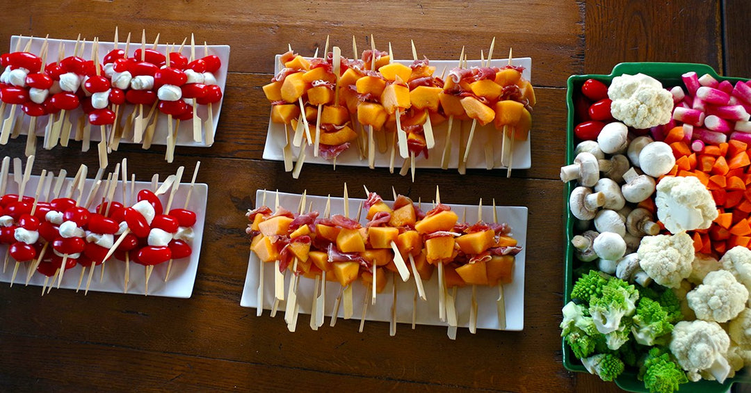 Appetizer skewers with tomatoes, mozzarella, melon and cured meat beside a tray filled with fresh vegetables.
