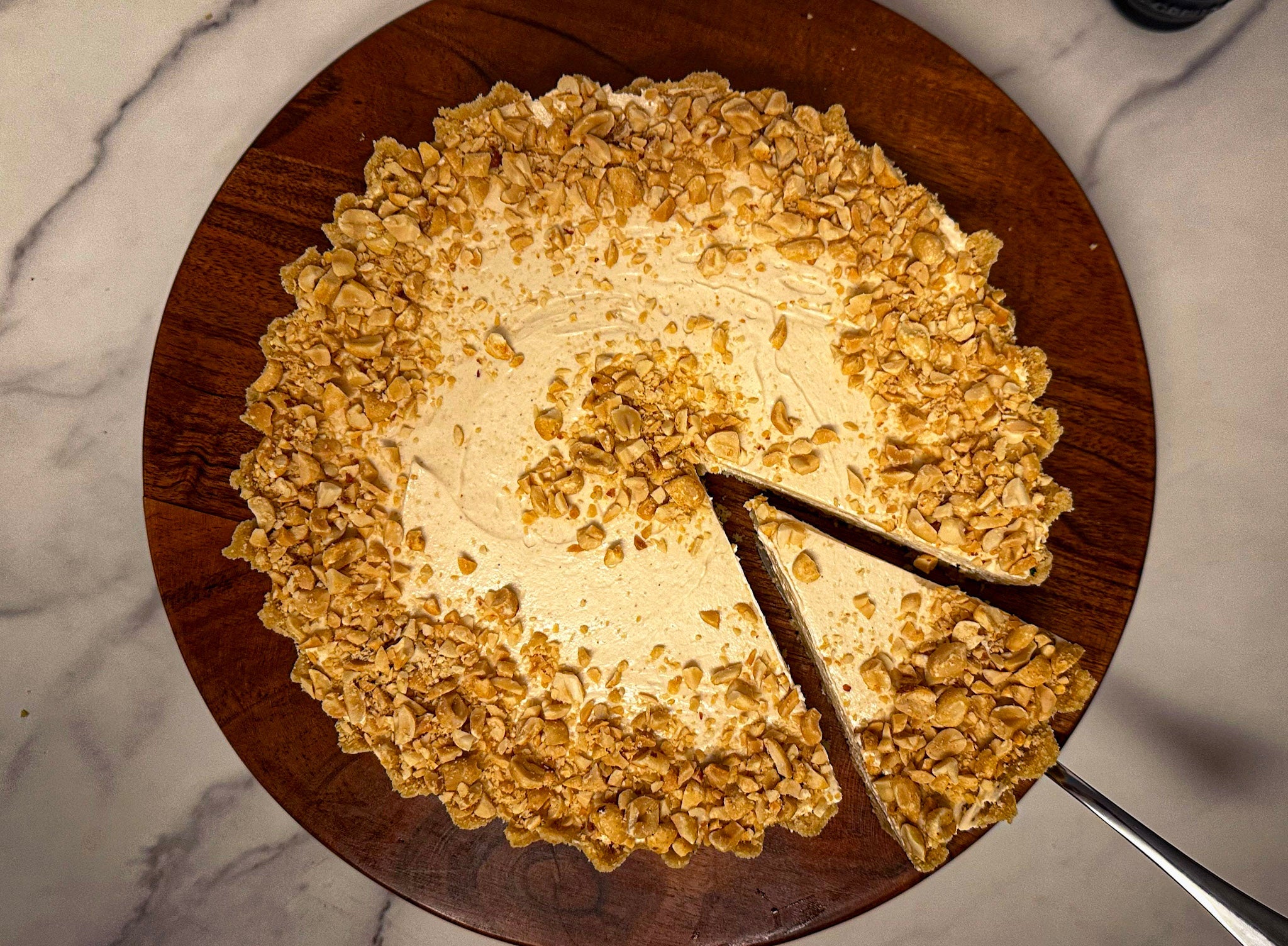 Peanut-topped cheesecake on a wooden board with one slice cut and lifted.