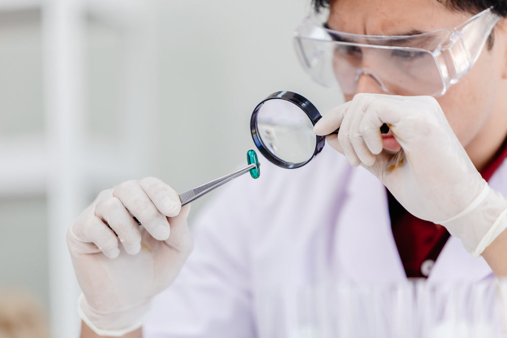 A scientist in a lab coat and safety goggles examines a green capsule with tweezers while using a magnifying glass, surrounded by laboratory equipment in a bright, modern setting.