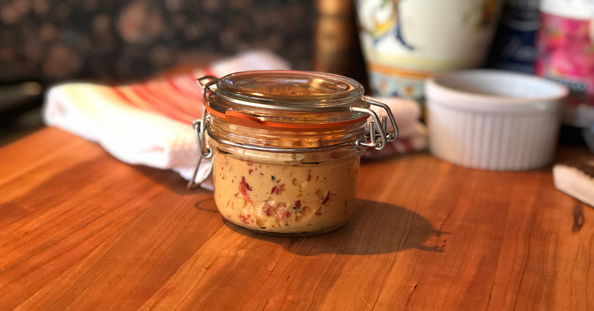 Small glass jar filled with creamy spread containing red bits, sitting on a wooden countertop.