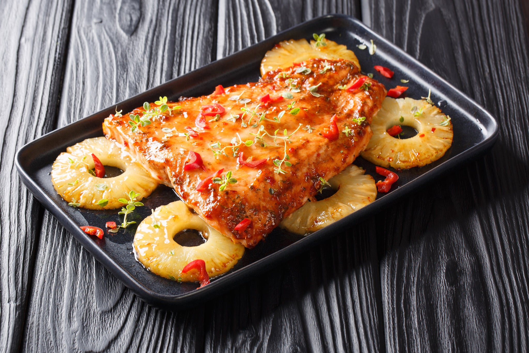 A glazed piece of fish is served on a black plate with pineapple rings and red pepper pieces, garnished with fresh herbs, against a textured wooden background.