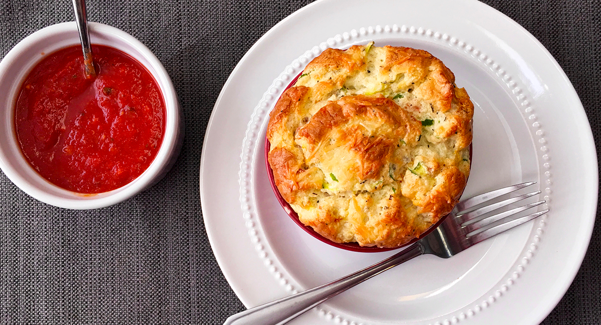 Individual savory soufflé in a ramekin on a white plate with a fork, served beside a small bowl of tomato sauce.