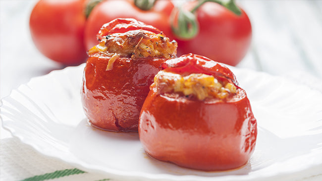 Two stuffed red bell peppers sit on a white plate, garnished with a golden filling, surrounded by fresh tomatoes in the background.