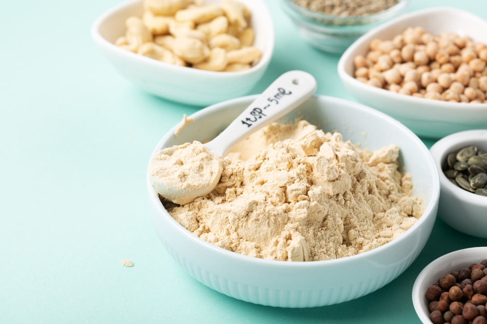 A bowl of light-colored vegan protein powder sits on a mint green surface, accompanied by various nuts and seeds in small bowls, highlighting ingredients for healthy shakes.