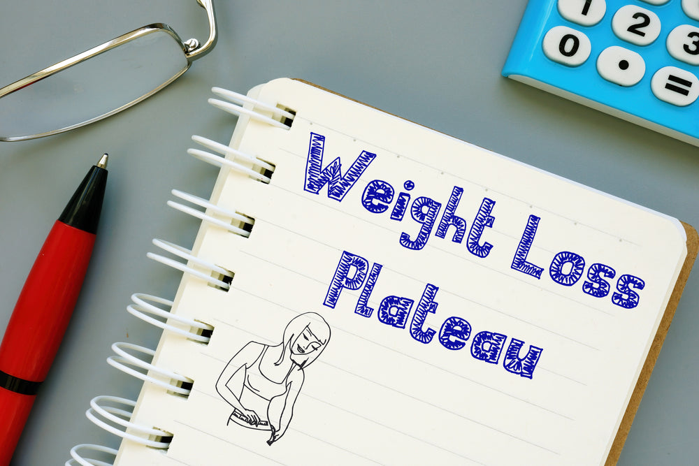 A notebook with the title "Weight Loss Plateau" is open, featuring a drawing of a woman measuring her waist, alongside a red pen and a blue calculator on a gray surface.