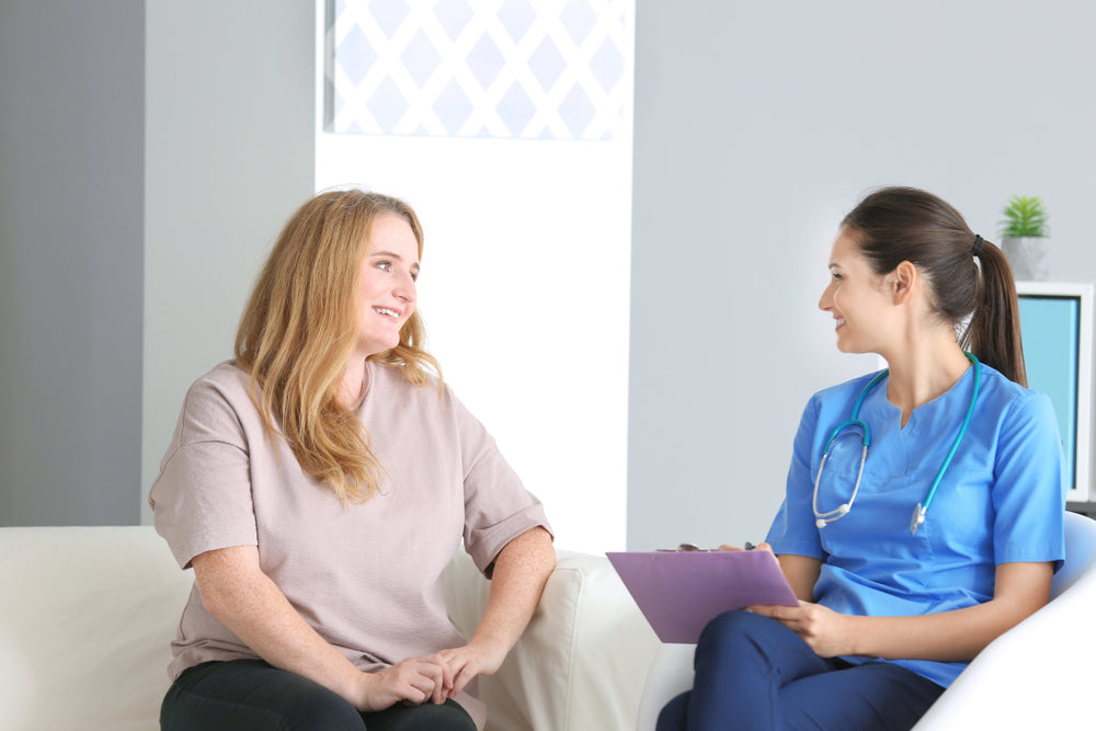 A woman with long hair smiles while sitting on a couch, talking to a nurse in scrubs who holds a clipboard, in a bright, modern office setting focused on post-bariatric surgery support.