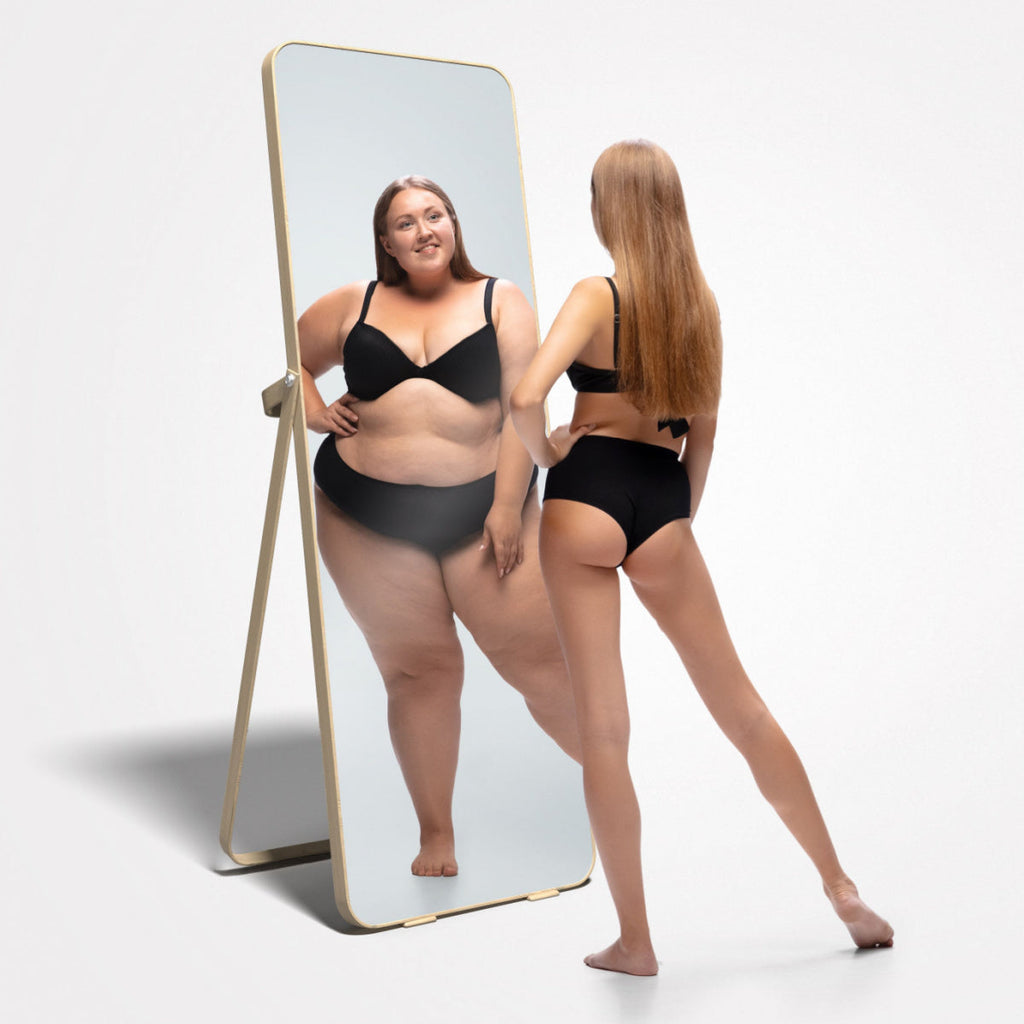 A woman in black underwear looks in a mirror that shows a larger-bodied reflection illustrating distorted self perception.