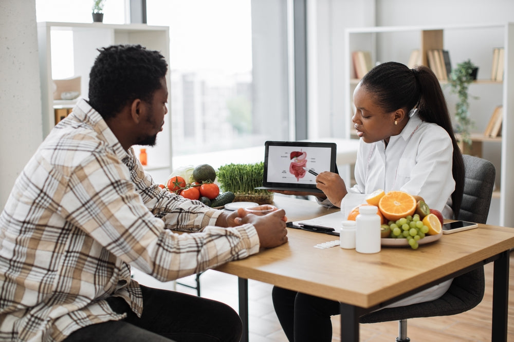 A healthcare professional in a white coat explains nutrition using a tablet to a man sitting at a table with fresh fruits and vegetables in a bright, modern kitchen setting.