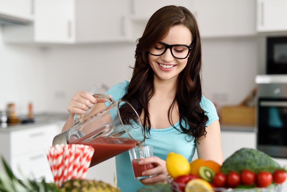A smiling woman in glasses pours a smoothie from a pitcher into a glass while surrounded by colorful fruits and vegetables in a bright kitchen.