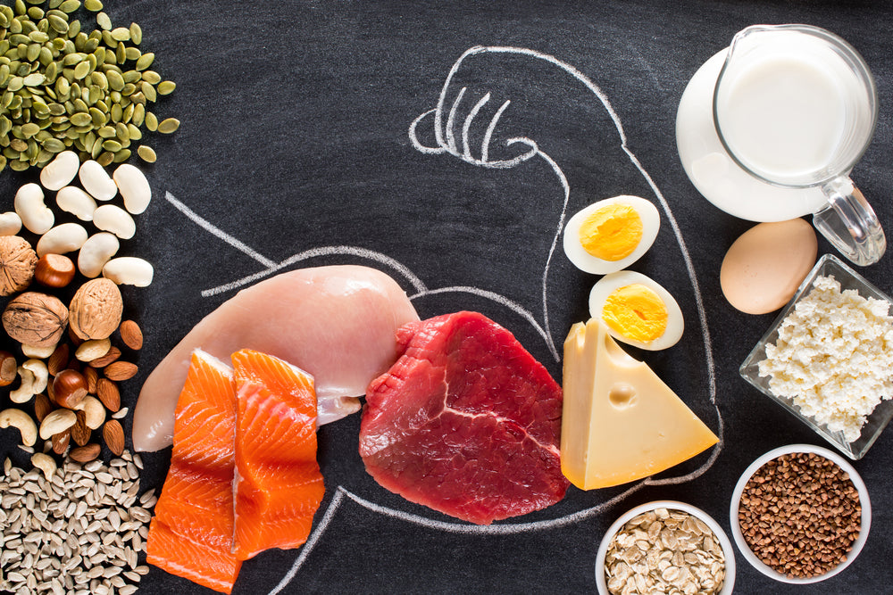 A variety of protein-rich foods, including meats, eggs, dairy, nuts, and seeds, are arranged around a chalk drawing of a flexing bicep on a black background, emphasizing their role in muscle building.