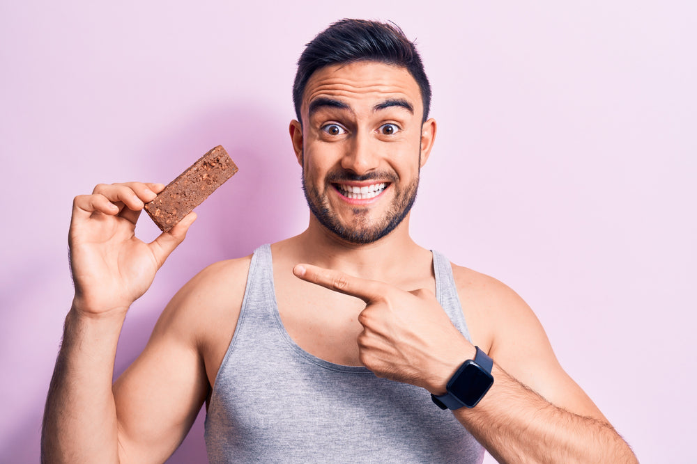 A smiling man in a gray tank top holds a chocolate bar in one hand and points at it with the other, against a light pink background.