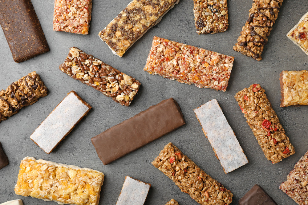 A variety of granola and protein bars in different flavors and textures are scattered across a gray surface, showcasing their colorful toppings and coatings.