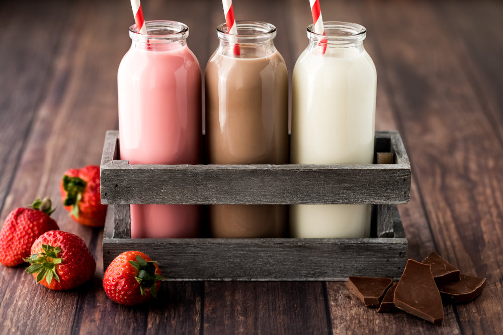 Three glass bottles of pink strawberry milk, chocolate milk, and white milk sit in a rustic wooden crate, surrounded by fresh strawberries and pieces of chocolate on a wooden table.
