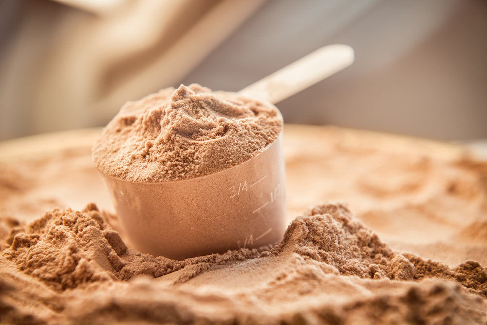 A brown powder fills a measuring cup, sitting atop a pile of similar powder on a wooden surface, with a blurred background suggesting a kitchen setting.