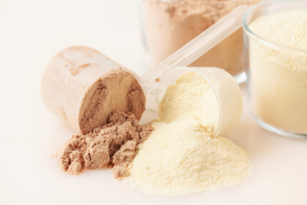 Two scoops of protein powder, one brown and one yellow, are spilled on a light surface with additional containers of powder in the background.