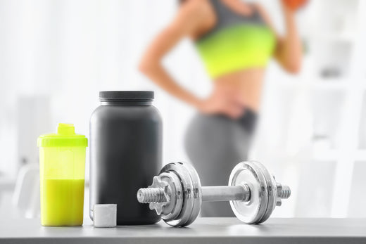 A black protein powder container and a yellow shaker bottle sit next to a silver dumbbell on a table, while a person in workout attire lifts a weight in the blurred background of a gym.