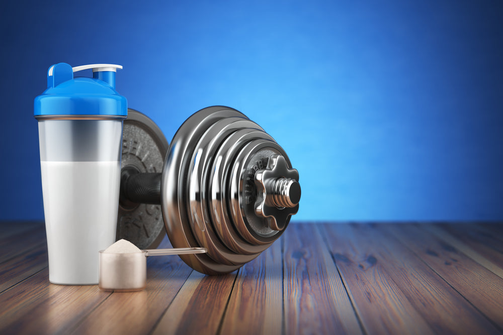 A protein shaker cup, a scoop of protein powder, and a shiny dumbbell are arranged on a wooden surface against a blue background, symbolizing fitness and nutrition for workout routines.