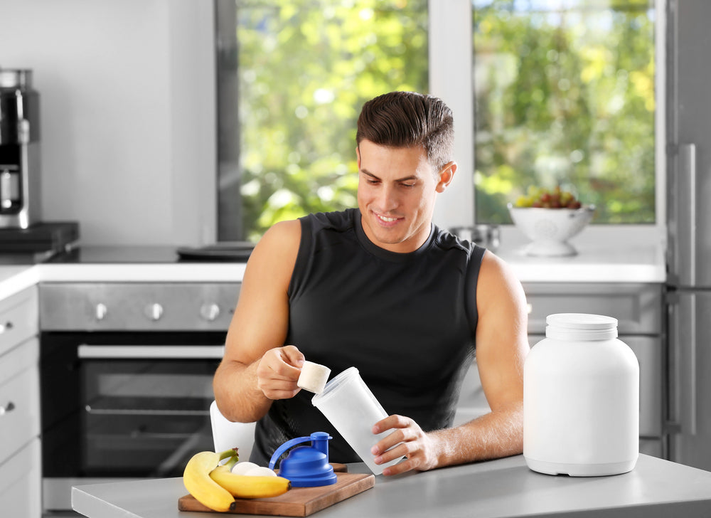 A man in a sleeveless shirt is smiling while measuring a scoop of protein powder into a white container on a kitchen counter, with bananas and a blue scoop nearby and greenery visible through the window.