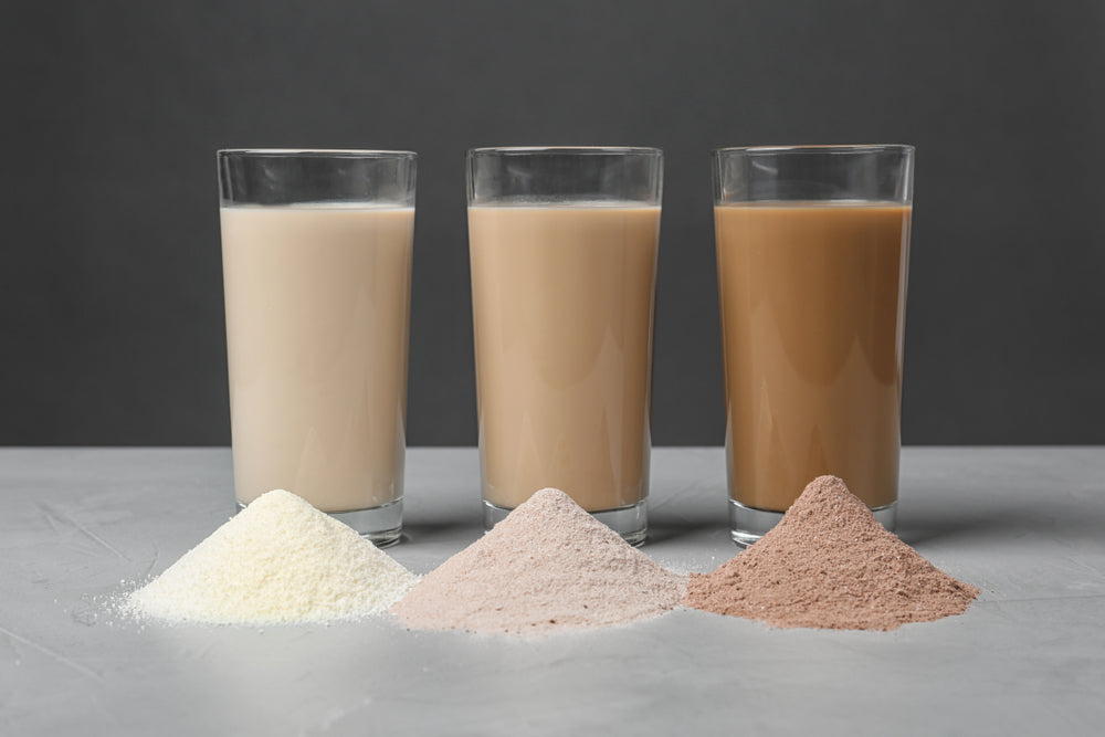 Three glasses of different colored drinks sit behind three mounds of powdered ingredients: white, pink, and brown, against a dark gray background.