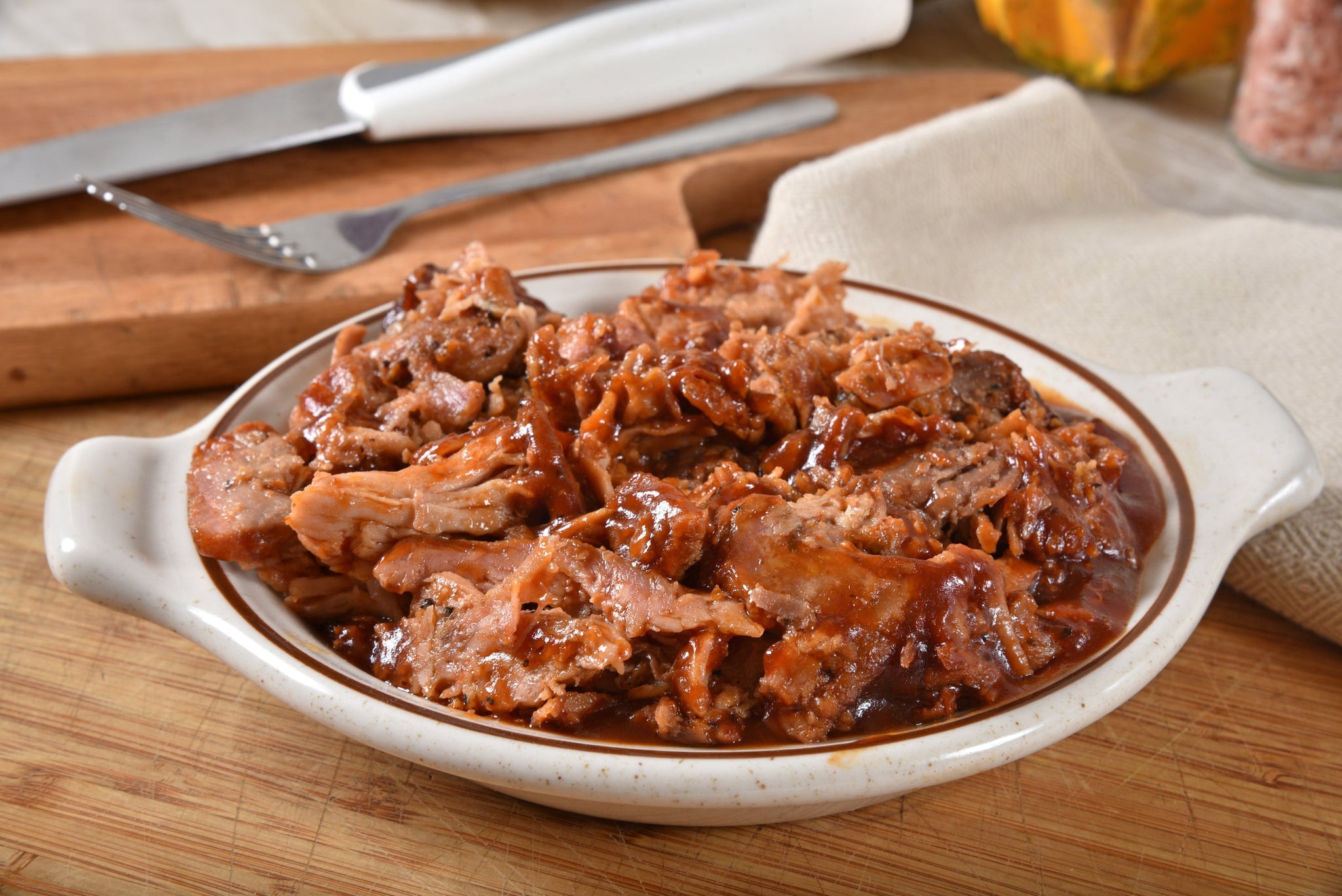 Dish of shredded, sauced pulled pork in a ceramic bowl on a wooden surface, with utensils and a napkin in the background.