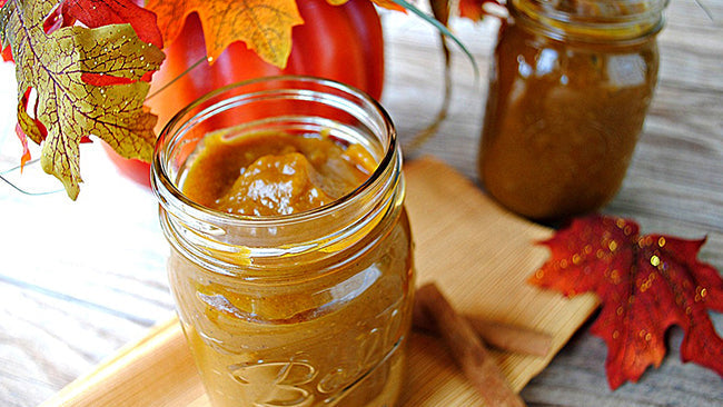 Jar of thick pumpkin butter on a wooden board, surrounded by autumn leaves and cinnamon sticks.