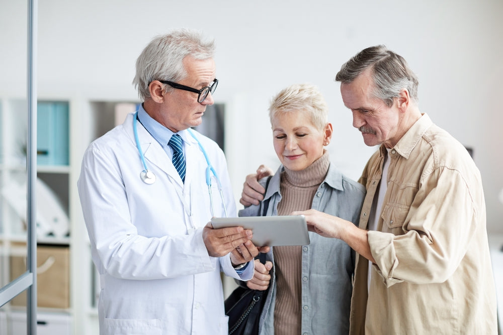 A doctor in a white coat shows a tablet to an older couple, who are smiling and engaged, in a bright, modern medical office setting.