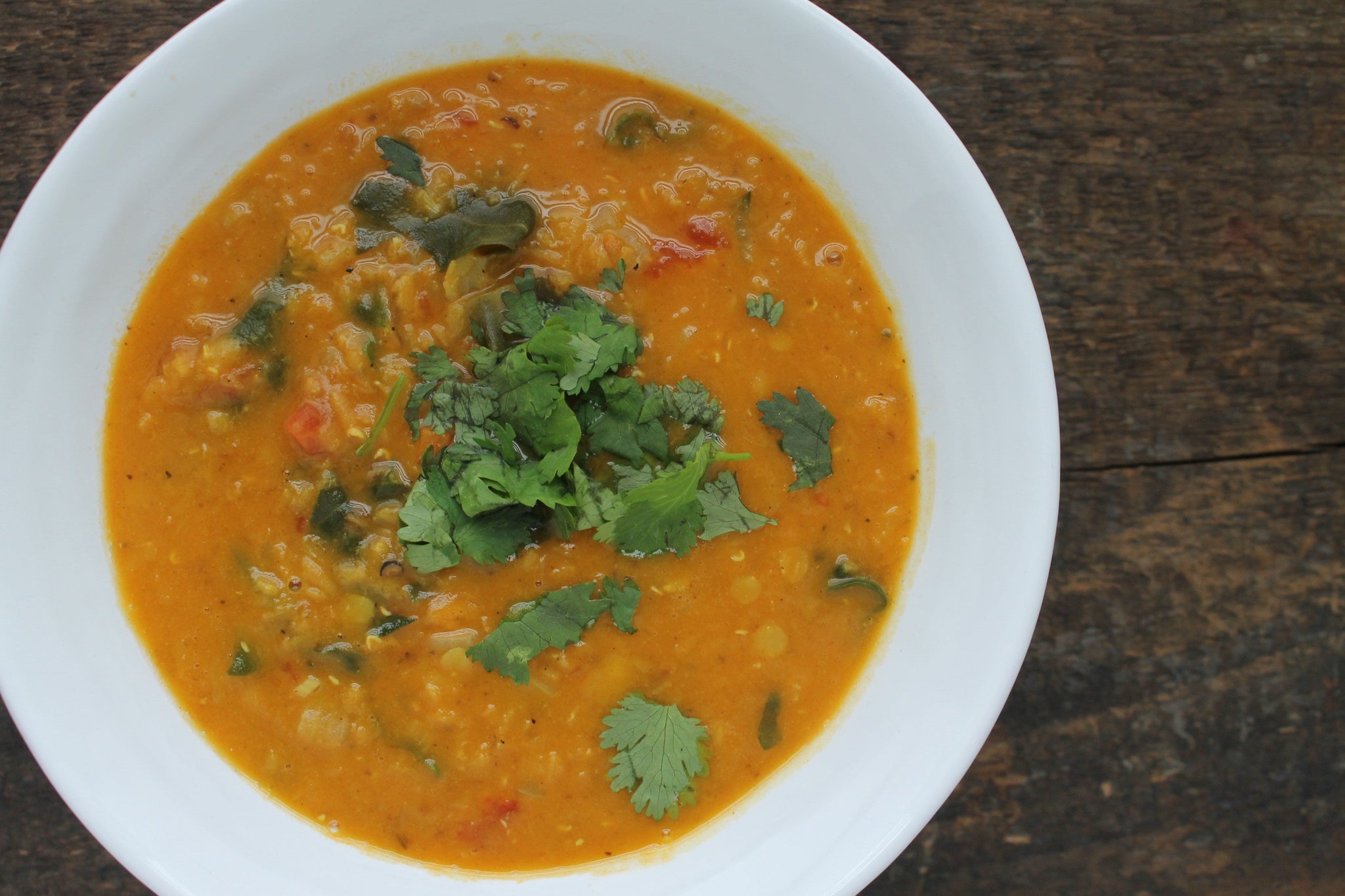 A bowl of orange lentil soup garnished with fresh cilantro, served in a white dish on a rustic wooden surface.