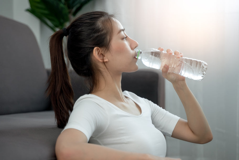 A woman with long hair is sitting on a couch, drinking from a clear water bottle, highlighting the importance of hydration for health and weight loss in a bright, indoor setting.