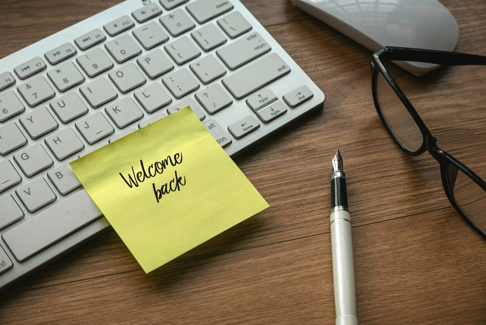 A yellow sticky note with "Welcome back" written on it is placed on a wooden desk next to a computer keyboard, a pen, and a pair of glasses.
