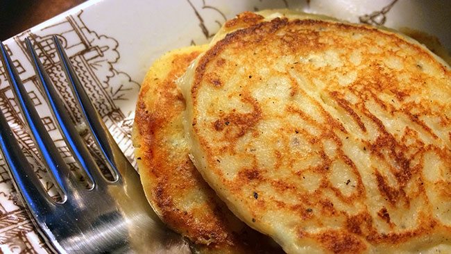 Two golden-brown pancakes are stacked on a decorative plate with a fork resting beside them, suggesting a cozy breakfast setting.