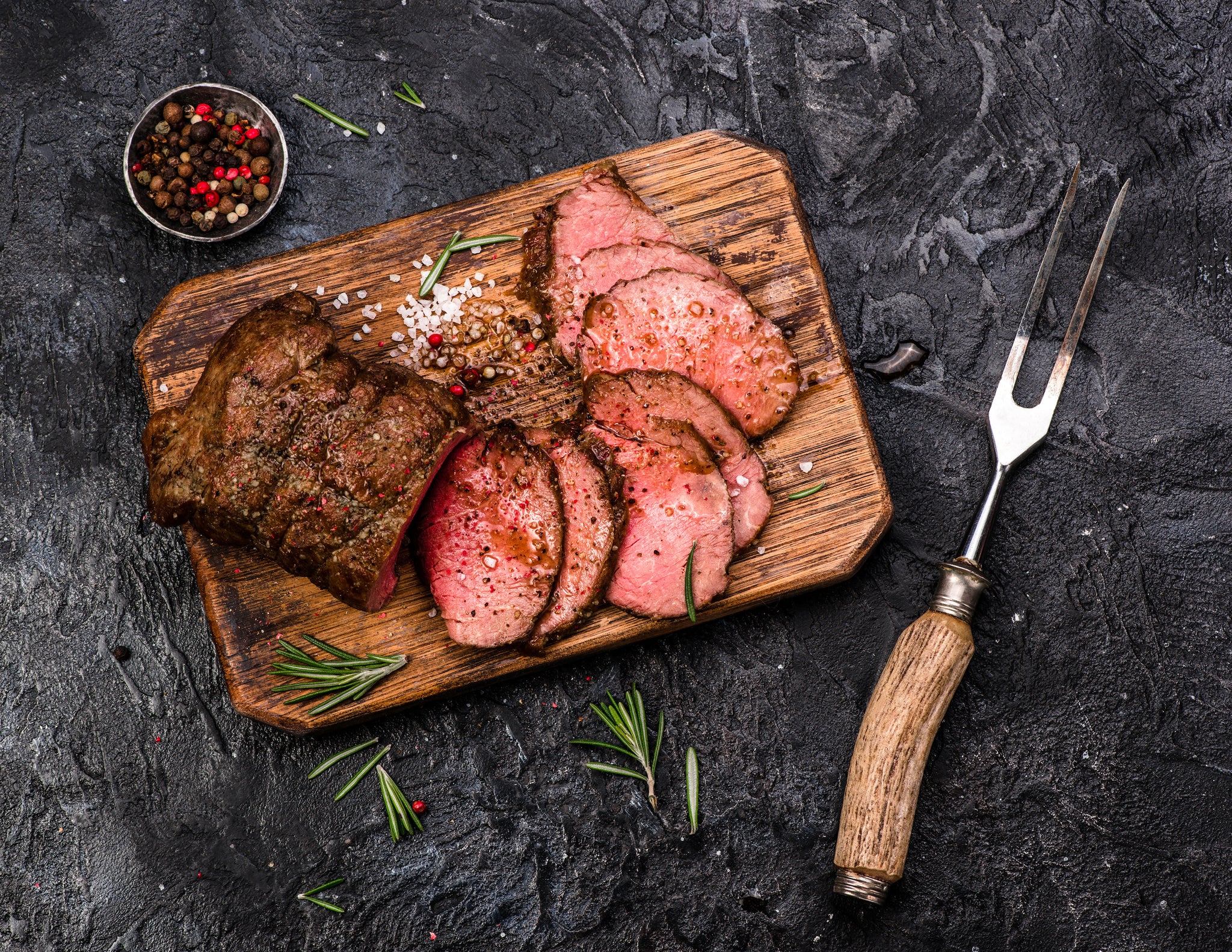 Sliced roast beef is arranged on a wooden cutting board, garnished with rosemary and coarse salt, alongside a metal fork and a small bowl of mixed peppercorns on a dark textured surface.