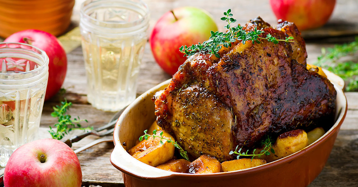 A roasted meat dish garnished with herbs sits in a brown baking dish, surrounded by cooked apples, with fresh apples and glasses of clear liquid on a rustic wooden table.
