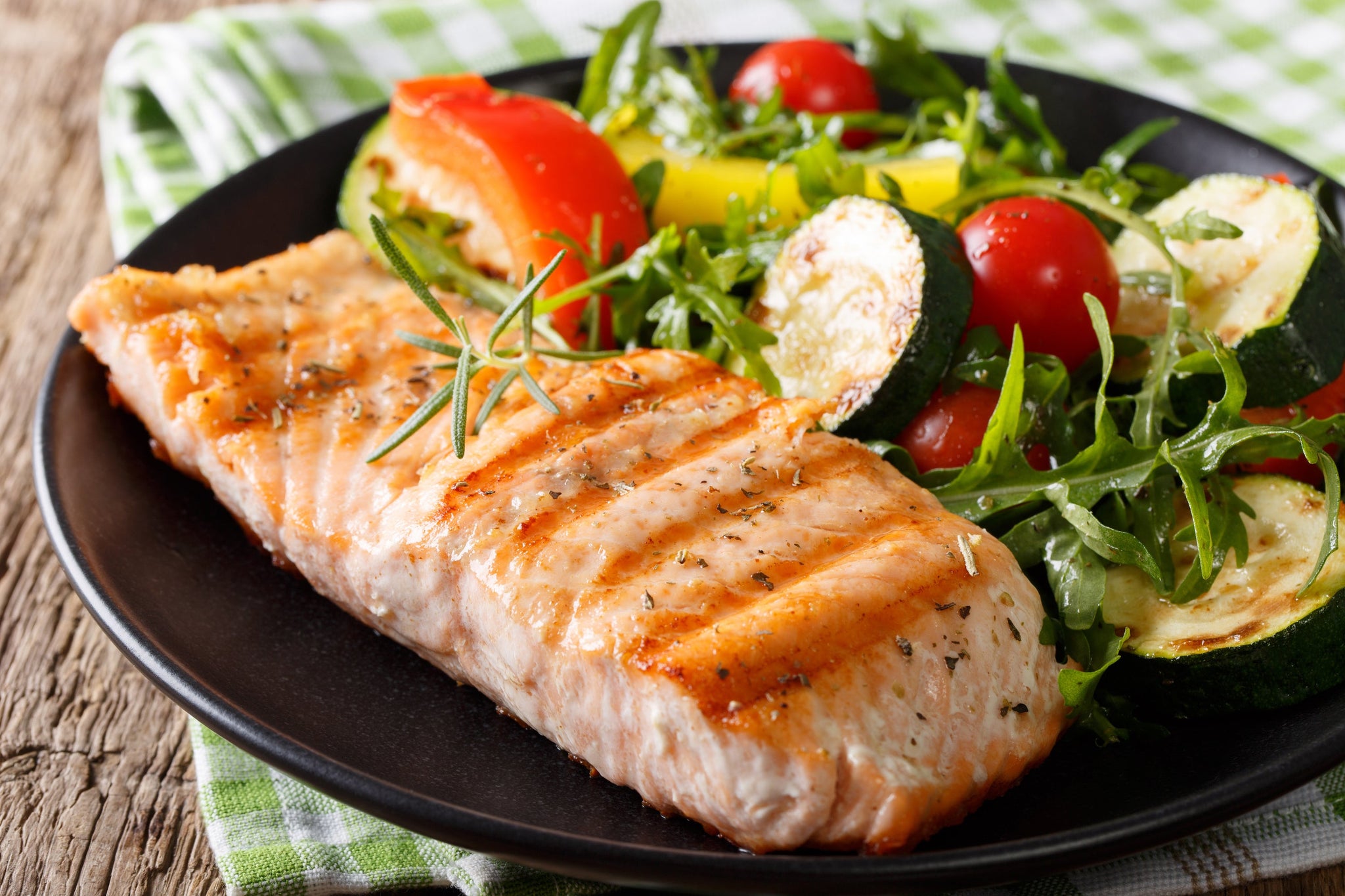 Grilled salmon fillet served on a plate with arugula, cherry tomatoes, zucchini, and roasted vegetables.
