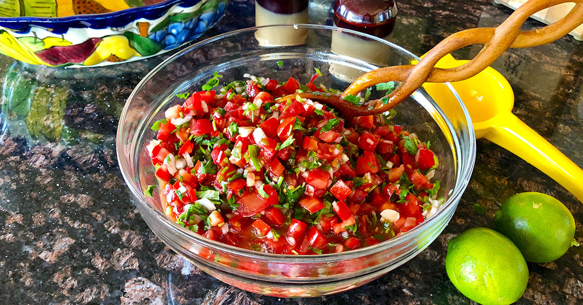 Glass bowl of fresh pico de gallo with tomatoes, onions, and cilantro, stirred with a wooden spoon beside limes.