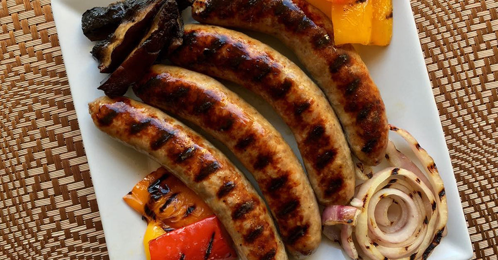 Grilled sausages served with charred peppers, onions, and mushrooms on a white rectangular plate.