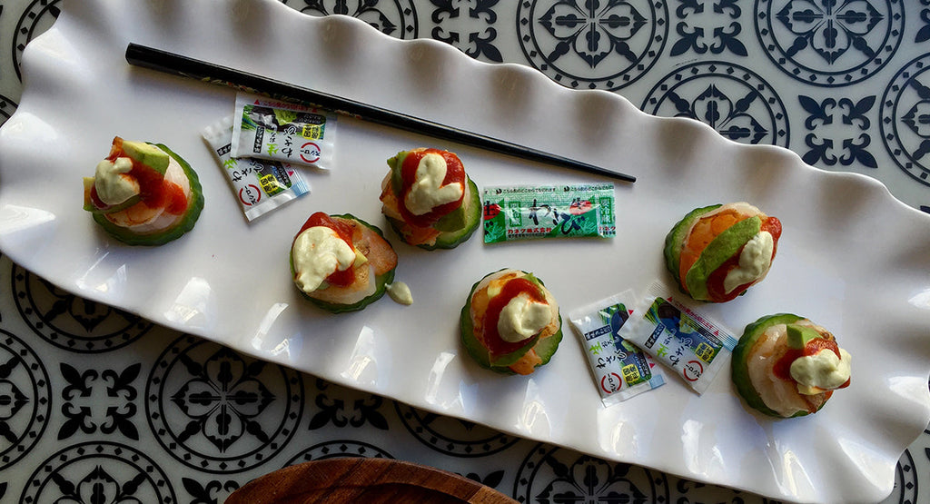 Small cucumber-based sushi bites topped with sauce, arranged on a white platter with wasabi packets and a chopstick.