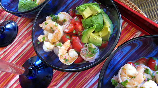 Three blue glasses filled with shrimp, diced tomatoes, and avocado, garnished with cilantro, are arranged on a colorful striped tablecloth, suggesting a fresh and vibrant seafood dish.