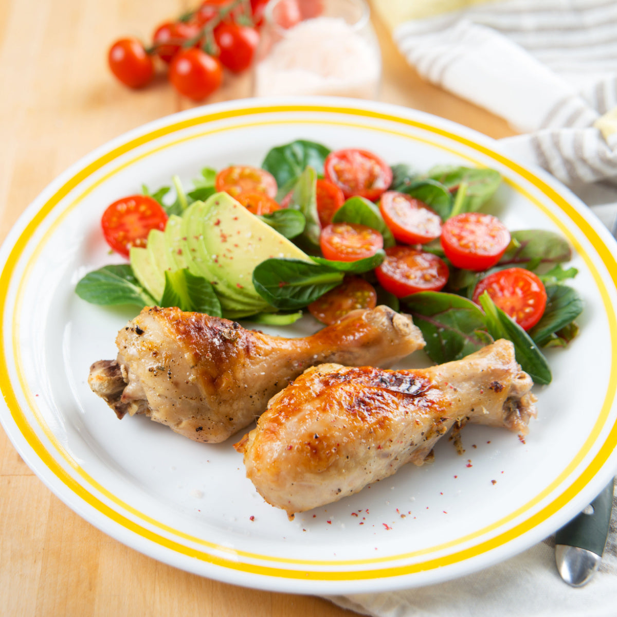 Two golden-brown chicken drumsticks are served on a white plate with a yellow rim, accompanied by a fresh salad of sliced tomatoes, avocado, and leafy greens, set on a wooden table.