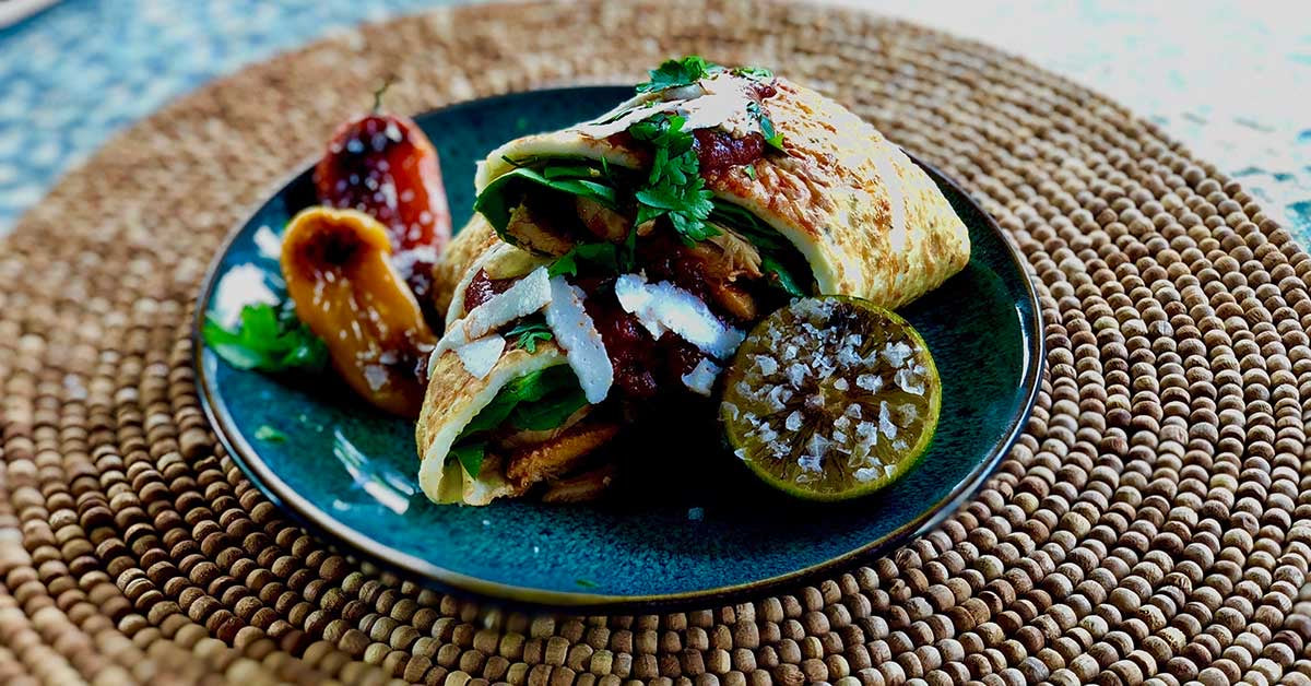 Plate of stuffed savory crepes with greens, sauce, grilled peppers, and a halved lime topped with salt.