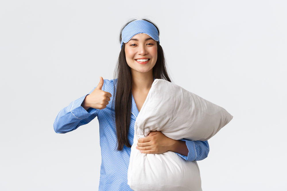 A young woman in blue pajamas and a sleep mask smiles and gives a thumbs-up while holding a fluffy white pillow against a plain background.