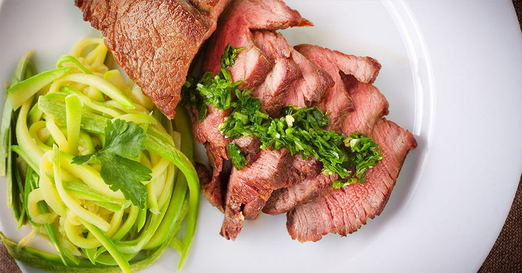 Slices of seared steak topped with green herb sauce, served beside zucchini noodles with parsley.