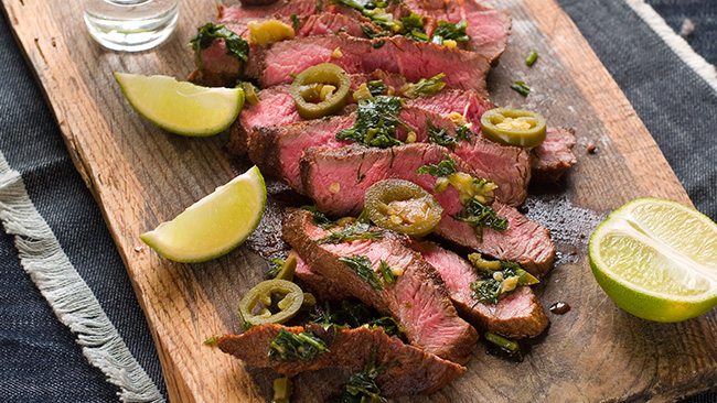 Slices of medium-rare steak topped with herbs and jalapeños on a wooden board with lime wedges.
